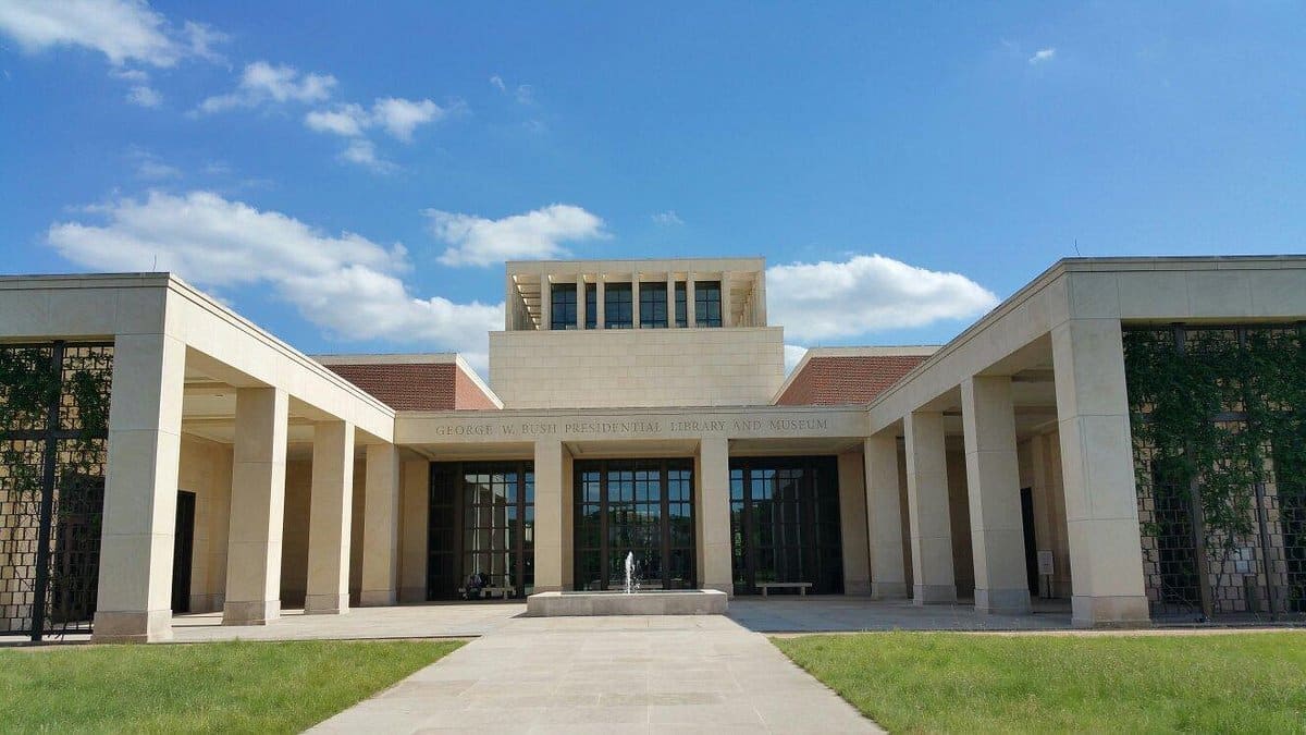 Front view of the George W. Bush Presidential Library and Museum with a blue sky and a few clouds in the background. The building has a modern design with large pillars, a central entrance, and well-managed visitor parking to accommodate guests efficiently.