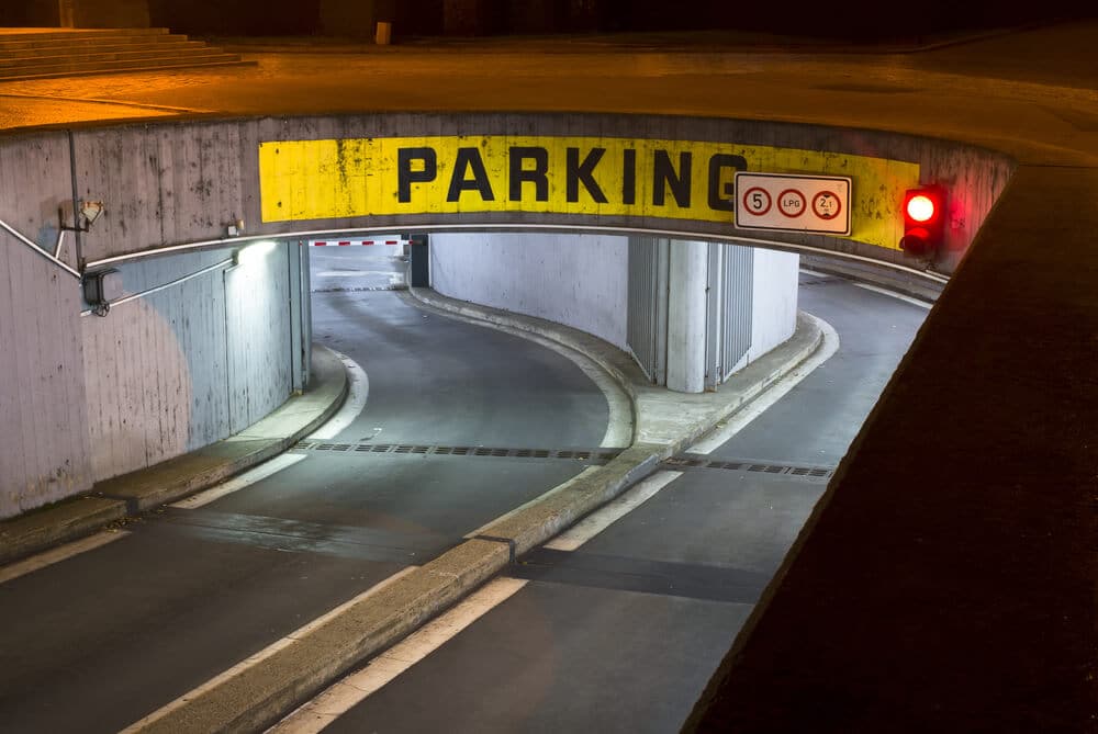 Entrance to a commercial parking garage at night, featuring a "Parking" sign, traffic signs, a barrier gate, and a red traffic light—a prime example of efficient parking lot management.