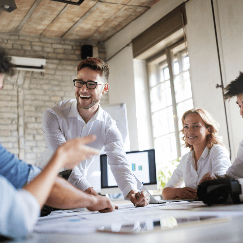 In a casual office setting, a group of people collaborates around a table with documents and a computer screen in the background. One person stands and smiles while others are seated and attentive. Just outside, effective parking management ensures visitor parking is hassle-free for all attendees.