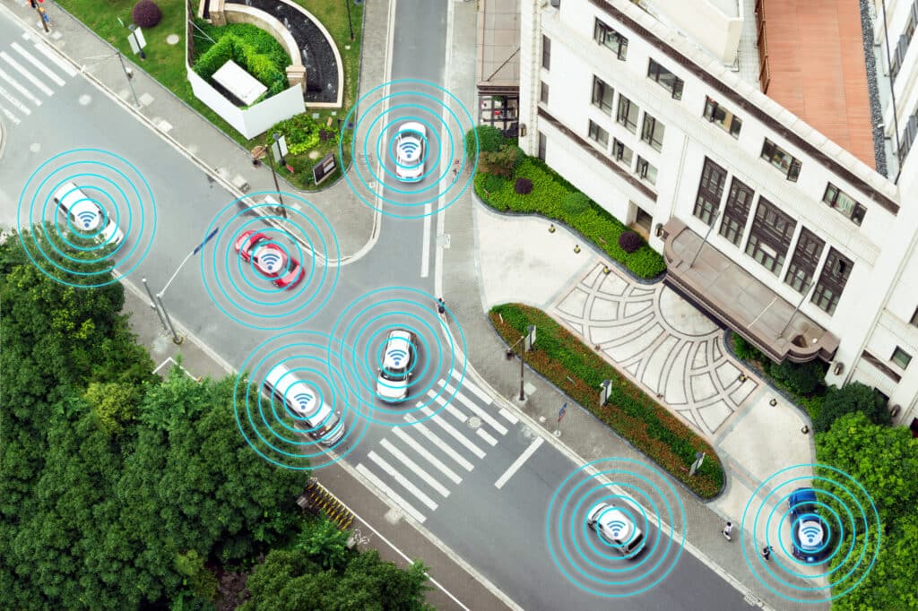 Aerial view of a city street with multiple cars showing connectivity signals around them, depicting a concept of connected or autonomous vehicles and innovative parking lot management systems.