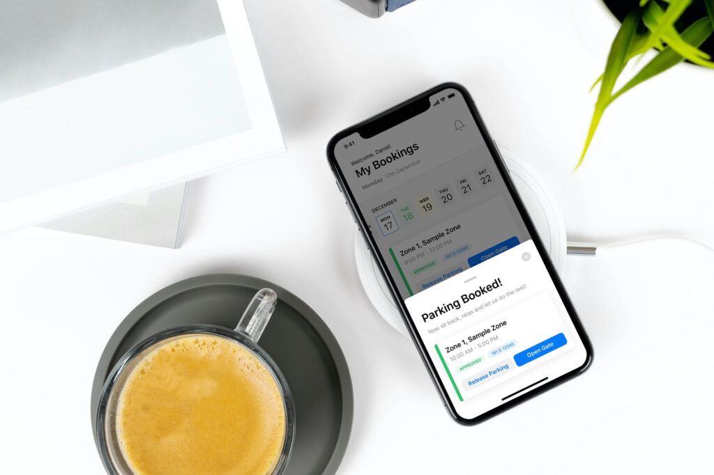 Smartphone on wireless charger displaying a Wayleadr parking booking confirmation on its screen. A cup of coffee sits beside the phone on a saucer. A portion of a table and plant are visible.