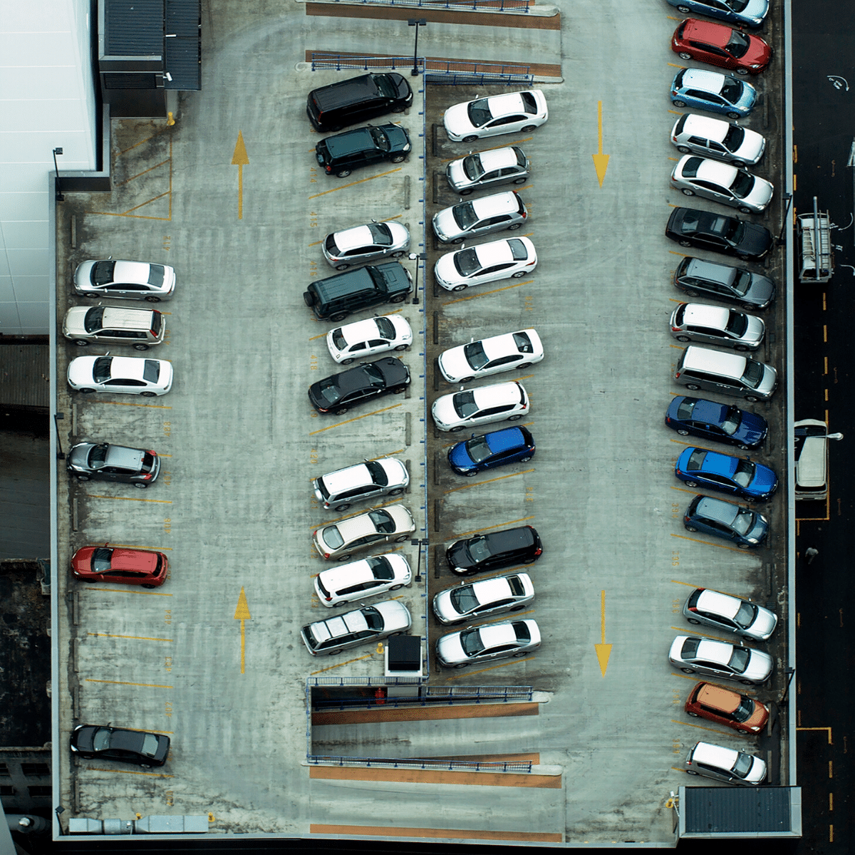 Aerial view of a commercial parking garage with multiple rows of parked cars, many in black, white, and various other colors, arranged in organized rows and columns. Wayleadr's parking lot management system ensures seamless organization and efficiency.