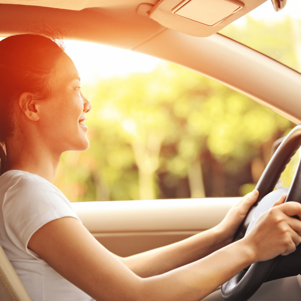 A woman is smiling and driving a car in a sunny, outdoor setting, effortlessly navigating through the visitor parking area.