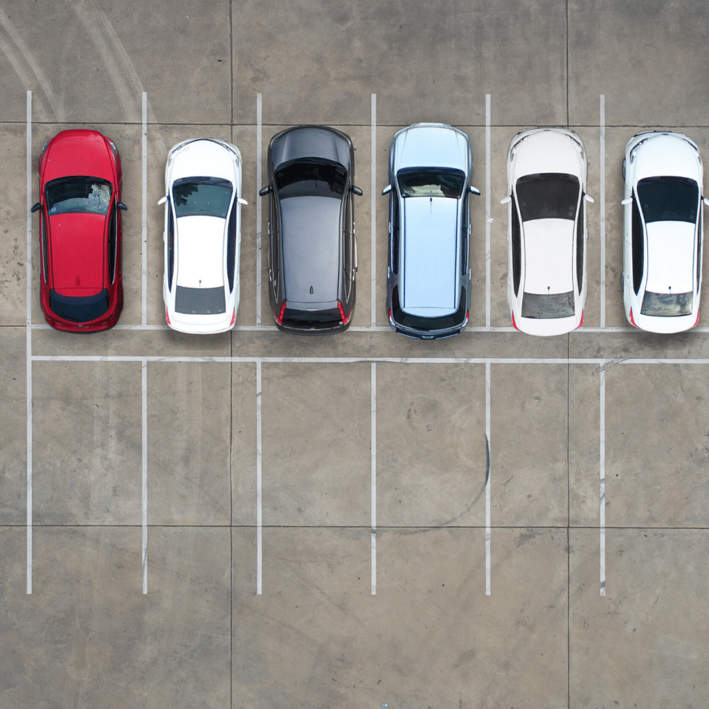 Aerial view of seven cars parked in a lot, with six spaces filled and one space on the far left vacant. The colors of the cars include red, white, gray, black, blue, and silver. This commercial parking setup showcases efficient parking management with Wayleadr's innovative technology.