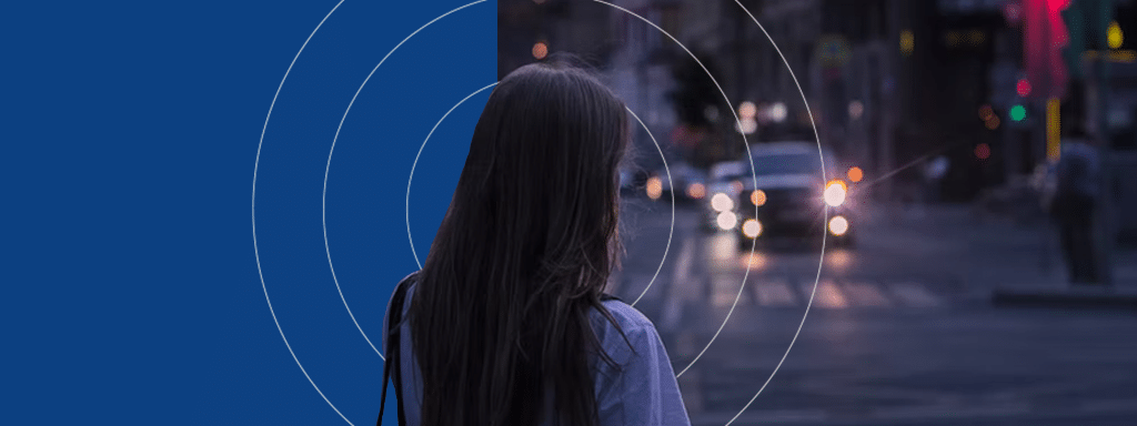 A person with long hair stands on a city street at dusk, facing away from the camera. Illuminated car headlights and streetlights are visible in the background, casting a glow that hints at a nearby parking lot system managing visitor parking efficiently.