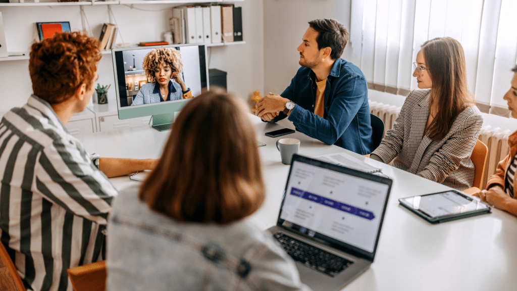 Four people are sitting around a table having a meeting, with one person joining via video call on a monitor. Laptops, notepads, and coffee cups are on the table as they discuss visitor parking and the advantages of using Wayleadr for efficient parking management.