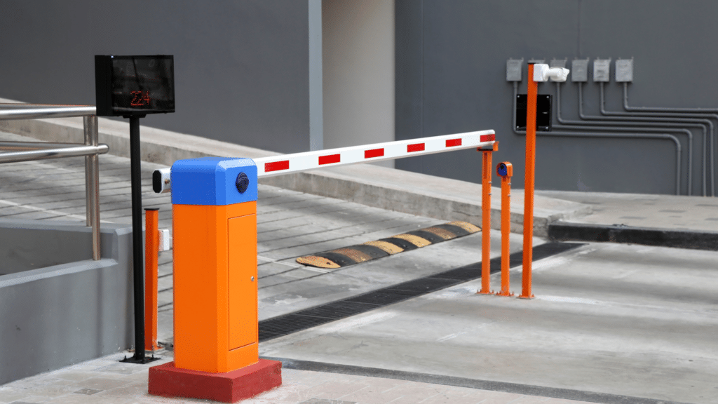 An automated parking barrier gate with orange and blue posts and a striped arm is positioned at the entrance of a parking facility, showcasing advanced parking lot systems. Nearby, a speed bump and electronic control panel are visible, ensuring seamless visitor parking management.