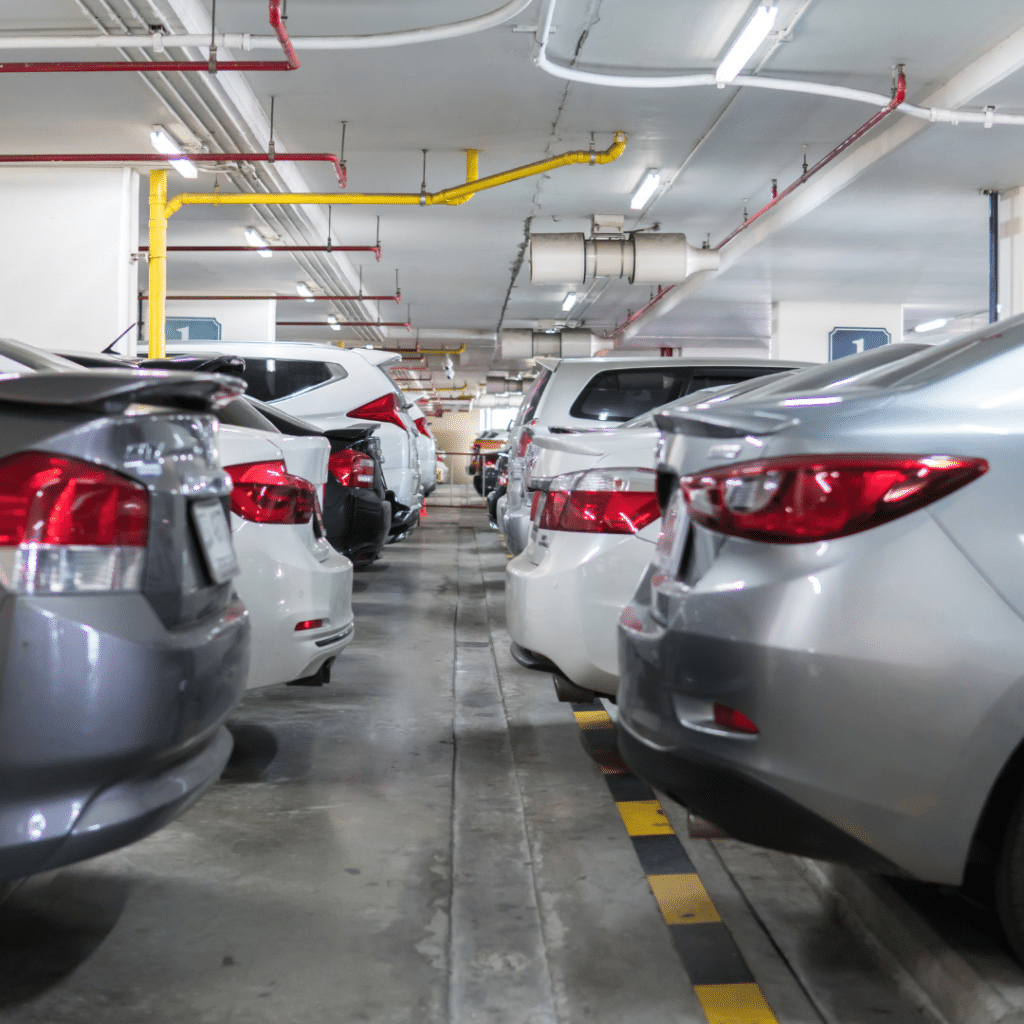 Underground commercial parking garage with rows of parked cars, illuminated by overhead fluorescent lights. The cars are parked closely together, filling most of the available spaces, demonstrating advanced parking lot management systems in place.
