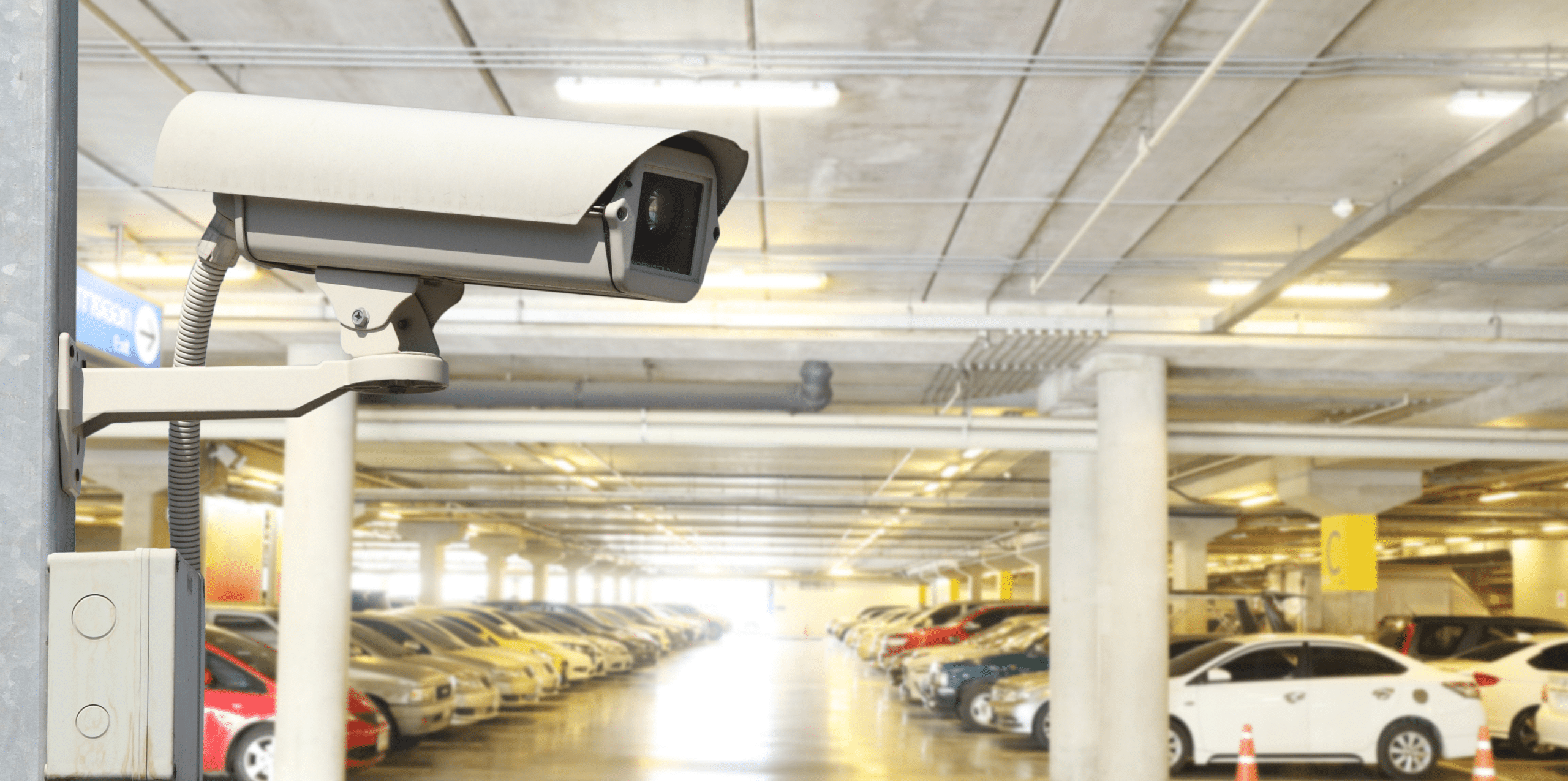 A surveillance camera is mounted on a wall, monitoring a well-lit visitor parking garage with numerous parked cars, enhancing parking lot management for Wayleadr.