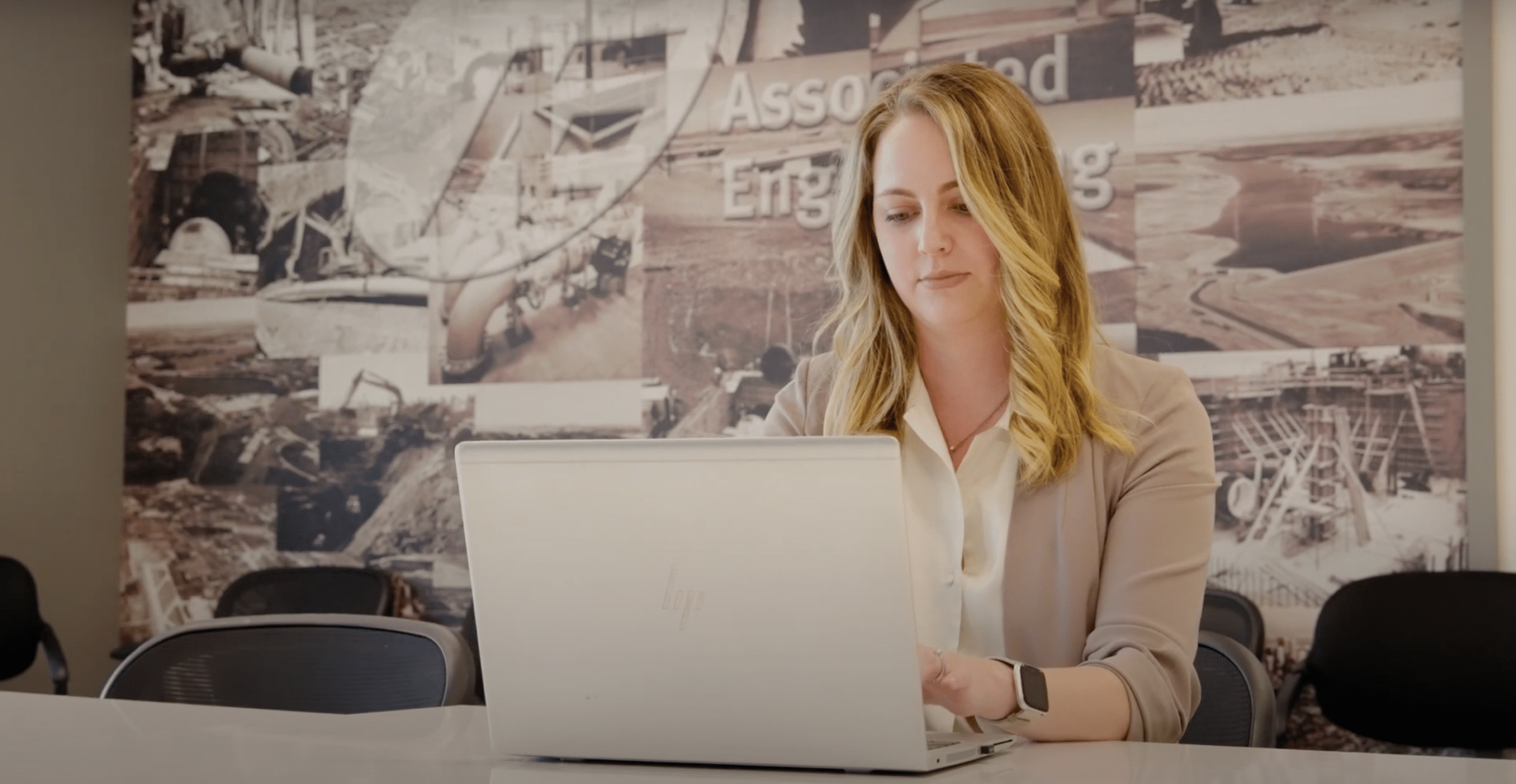 A woman with blonde hair uses a laptop while seated at a table in a conference room with an industrial-themed mural, likely reviewing Wayleadr's parking lot management system.