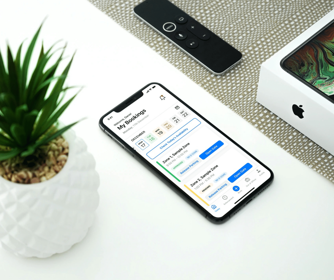 A smartphone displaying a bookings app sits on a table next to a potted plant, a remote control, and an iPhone box, seamlessly integrating with Wayleadr's parking management system.