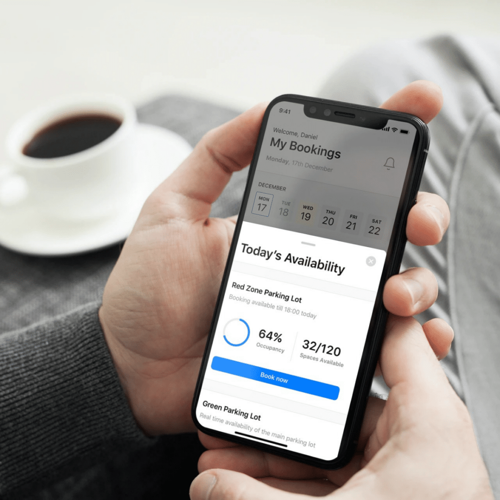 A person holds a smartphone displaying a Wayleadr app, showing commercial parking statistics with 64% occupancy and 32 out of 120 spots available in the Red Zone Parking Lot. In the background, a coffee cup rests on a saucer.