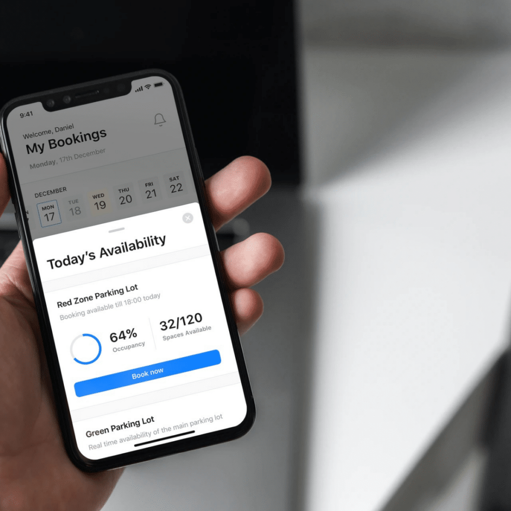 A hand holding a smartphone displaying a parking reservation app shows today's availability for visitor parking. The Red Zone Parking Lot is 64% occupied, and 32 out of 120 spaces are available, making it an effective tool for efficient parking lot management.