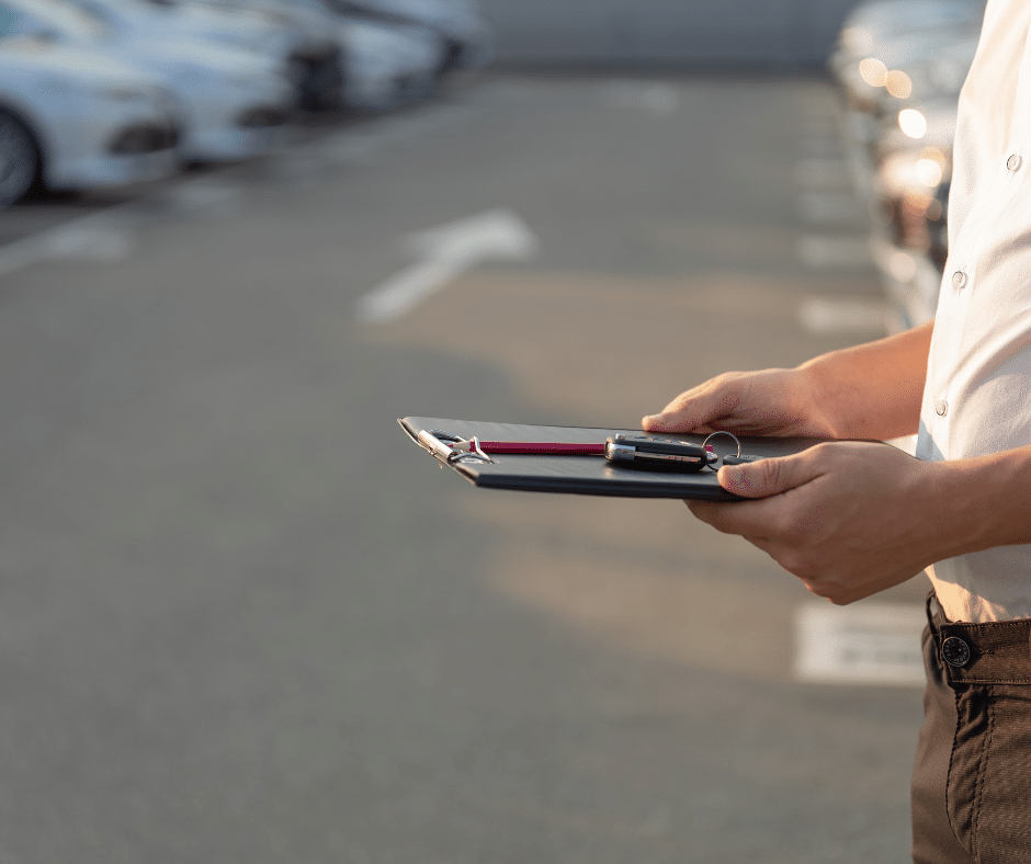 Person holding a clipboard and pen in a parking lot, with rows of parked cars in the background, overseeing Wayleadr's innovative parking lot management system.