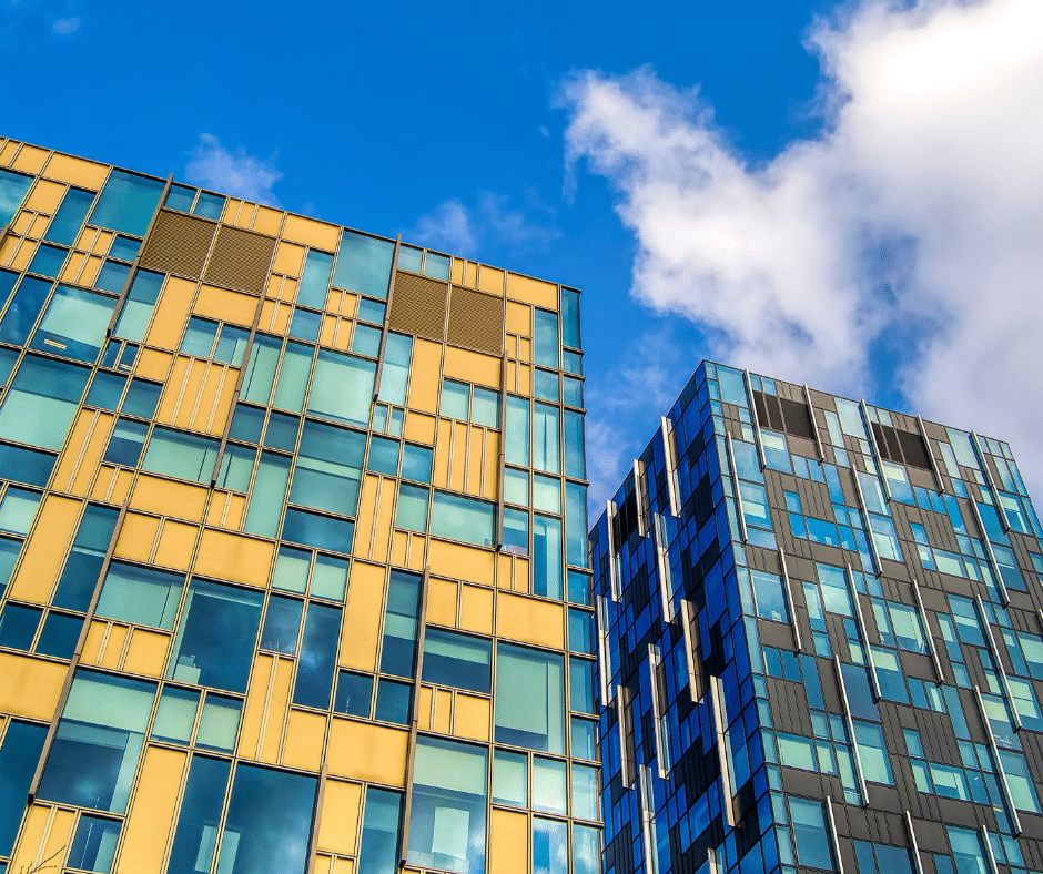 Two modern, geometric-patterned buildings with blue and yellow panes stand against a bright blue sky with scattered clouds. Below, a well-organized visitor parking system managed by Wayleadr ensures convenience for all guests.