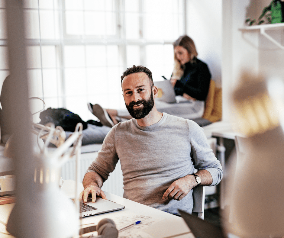 A bearded man sitting at a desk and using a laptop smiles at the camera. A woman sits in the background by a window, working on her laptop, possibly managing commercial parking solutions with Wayleadr.