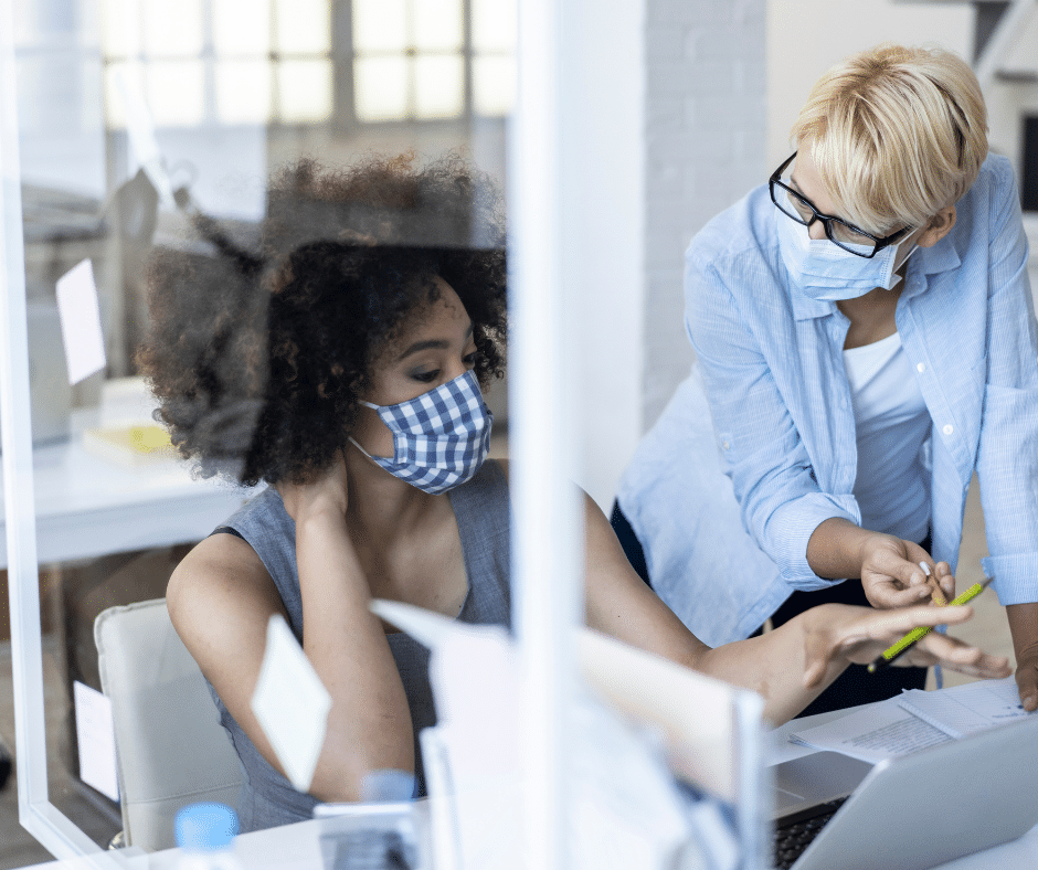 Two individuals wearing masks, one seated and the other standing, are discussing something over an open laptop in an office setting, possibly reviewing commercial parking data.