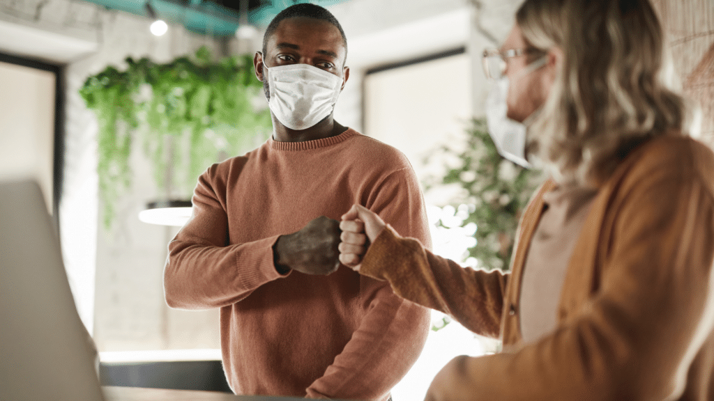 Two individuals wearing masks engage in a fist bump. One is a man with short hair, and the other is a person with long hair and glasses. They are standing near a visitor parking area in an indoor setting with plants in the background.
