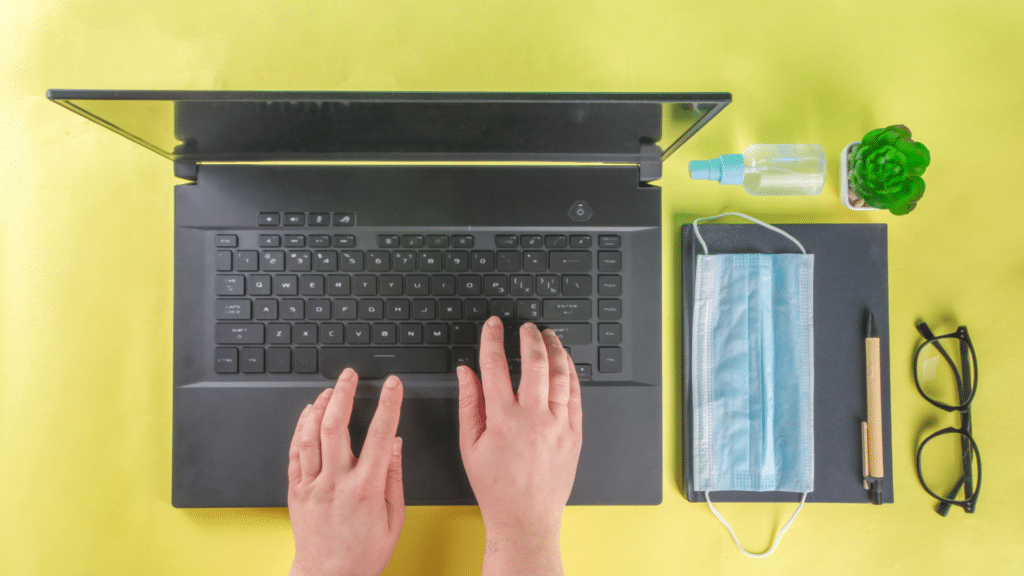 A person types on a laptop with a face mask, notebook, pen, hand sanitizer, glasses, and a small plant nearby on a yellow surface, efficiently working on parking management solutions.