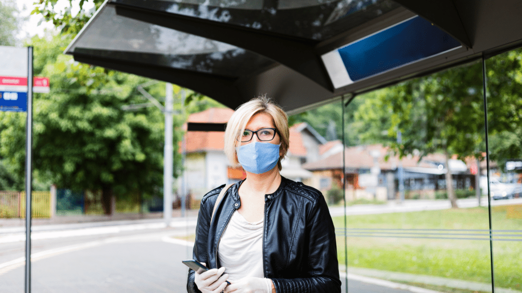 A person wearing a face mask and gloves stands at a bus stop while holding a smartphone, perhaps checking their Wayleadr app. Trees and buildings are visible in the background.