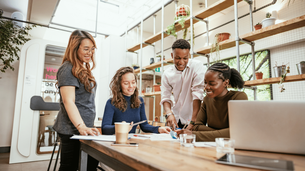 Four people are gathered around a table in a modern office, collaborating on a project. There are papers, pens, and a coffee cup on the wooden table, with plants and shelves in the background. The discussion is centered around using Wayleadr for innovative parking management solutions in their commercial space.