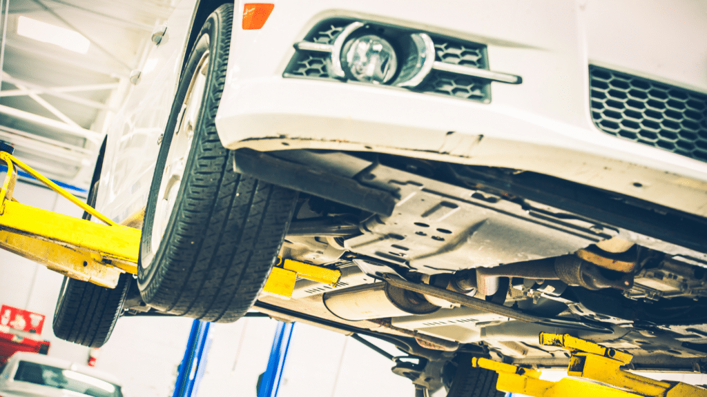 A white car is lifted on a yellow hydraulic lift in a mechanic's workshop, showing its undercarriage and front wheel. The advanced parking lot systems ensure that vehicles are efficiently organized before they even reach the workshop.