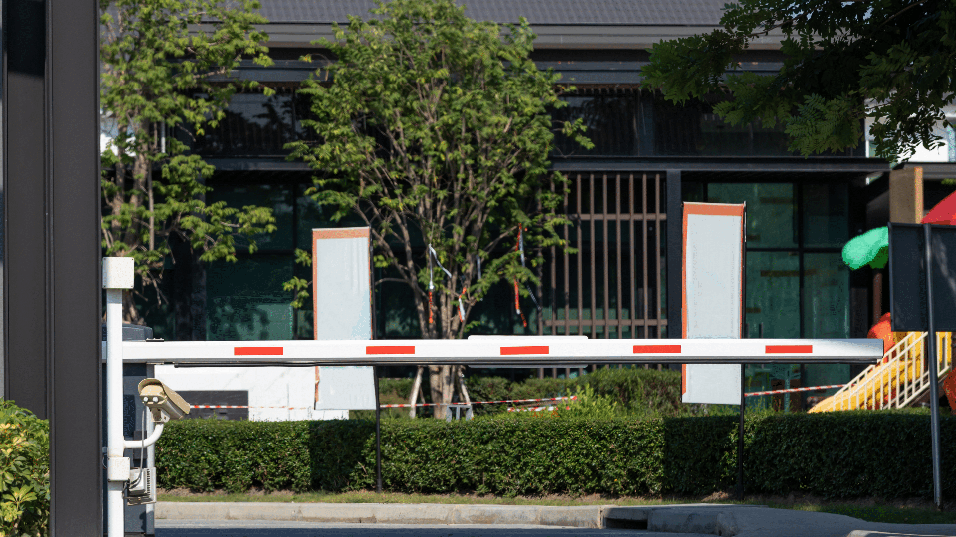 A closed parking gate with a red and white barrier arm blocks the entrance to the visitor parking lot in front of a building. Trees and a playground slide are visible in the background, showcasing effective parking management.