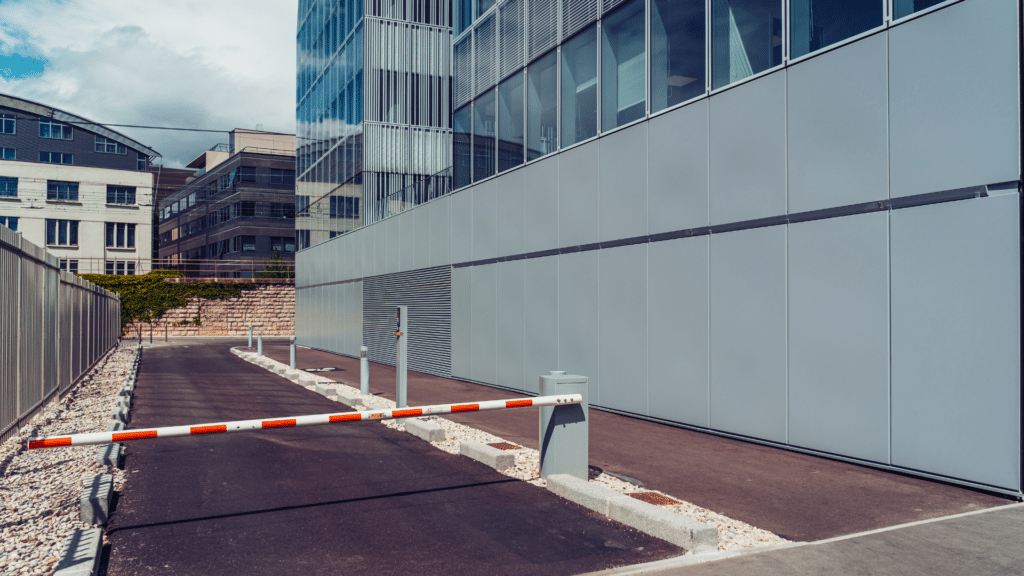 A closed white and red boom barrier on a paved narrow road adjacent to a modern glass and metal building on a clear day, exemplifying the efficiency of Wayleadr's parking lot management solutions.