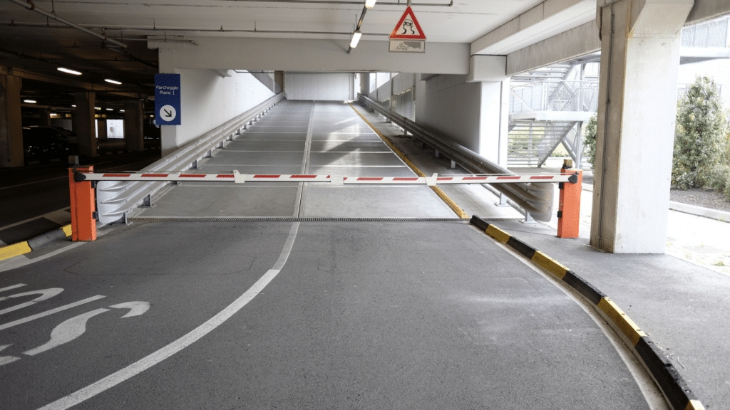 A vehicle entrance ramp in a parking garage features a lowered red and white barrier gate and speed limit markings on the ground. Signs provide directions and parking information, demonstrating efficient commercial parking management.