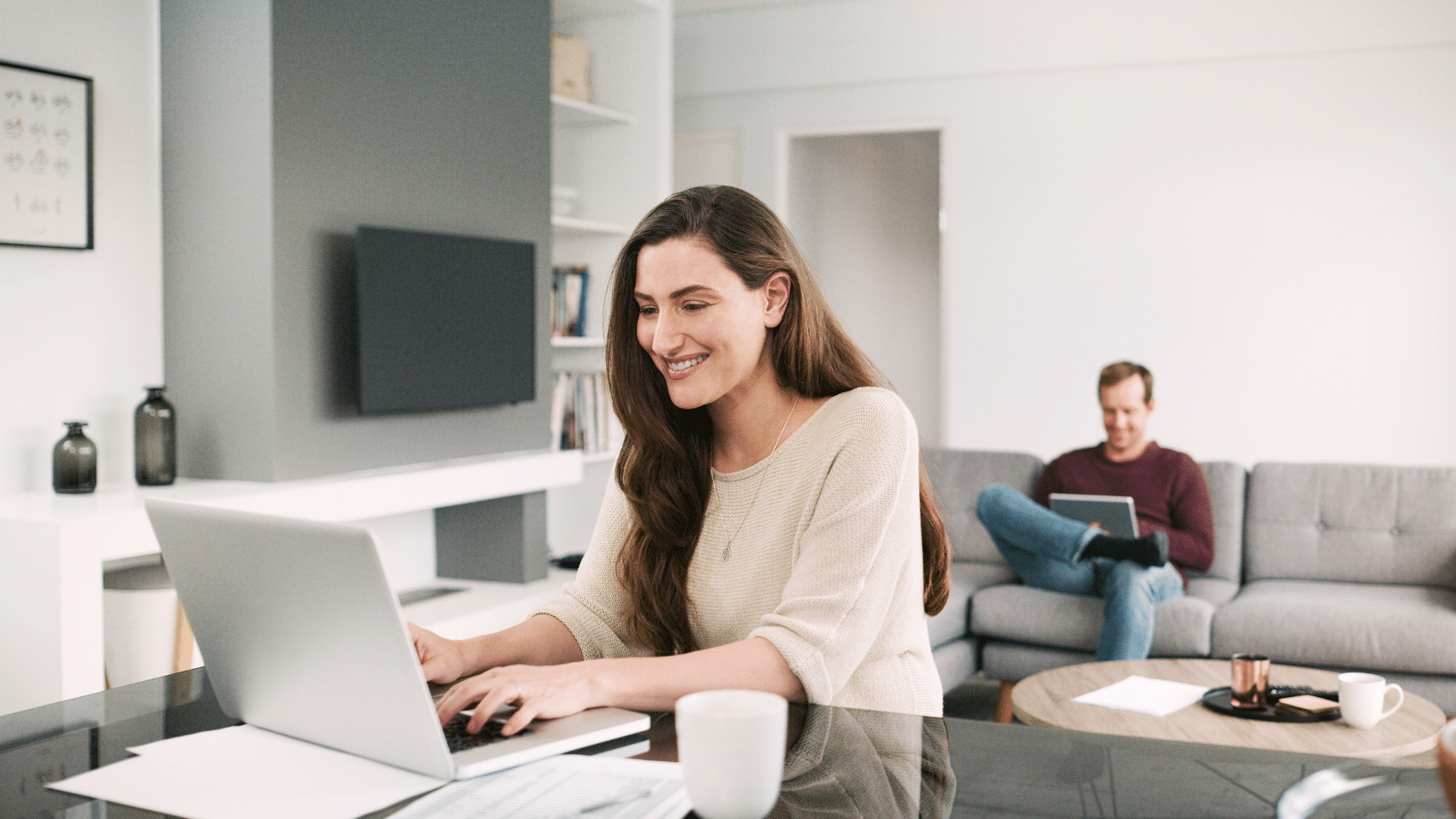 A woman is using a laptop at a table in a modern living room, possibly researching parking lot management systems, while a man sits on a couch in the background with a tablet.