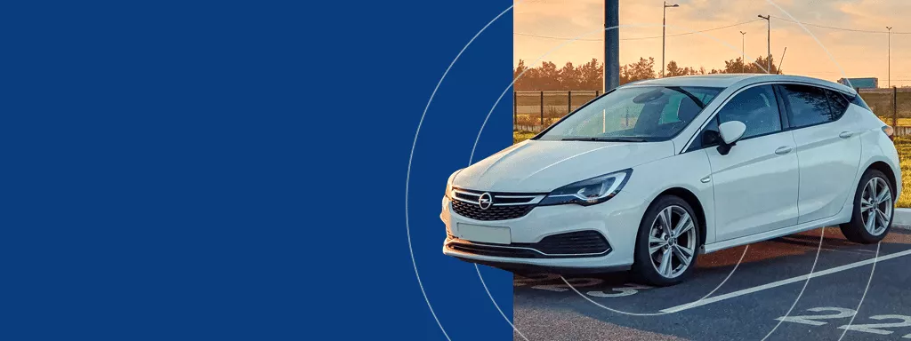 White Opel hatchback parked in a commercial parking lot at sunset, shown partially against a blue background.