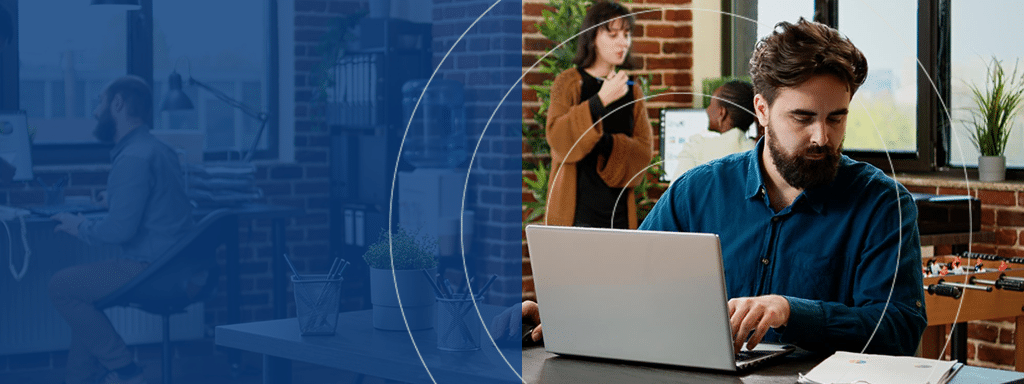A man with a beard works on a laptop at a desk. In the background, a woman and another person discuss parking lot management near a whiteboard in a modern office with brick walls and large windows.