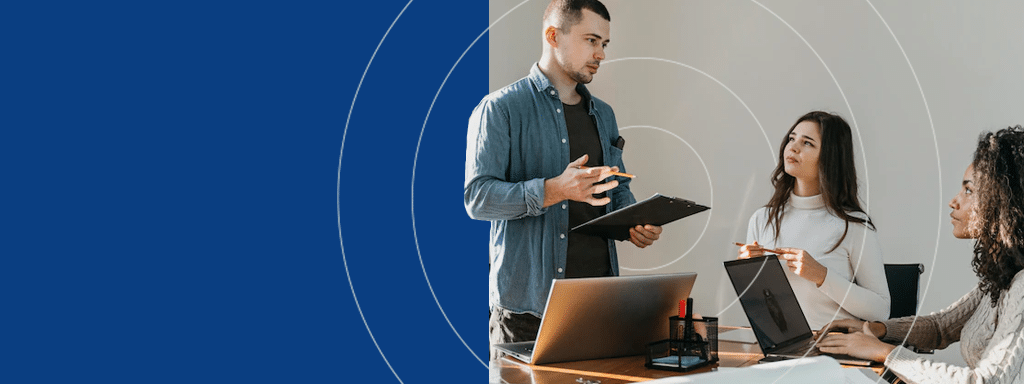 A man stands holding a clipboard, discussing parking management with two women seated at a table with laptops. They are engaged in conversation about optimizing visitor parking in a modern office setting.