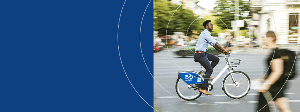 A man is riding a bike-sharing bicycle in an urban area, while commercial parking spaces managed by Wayleadr are visible in the background. A person walking by is blurred in the foreground.