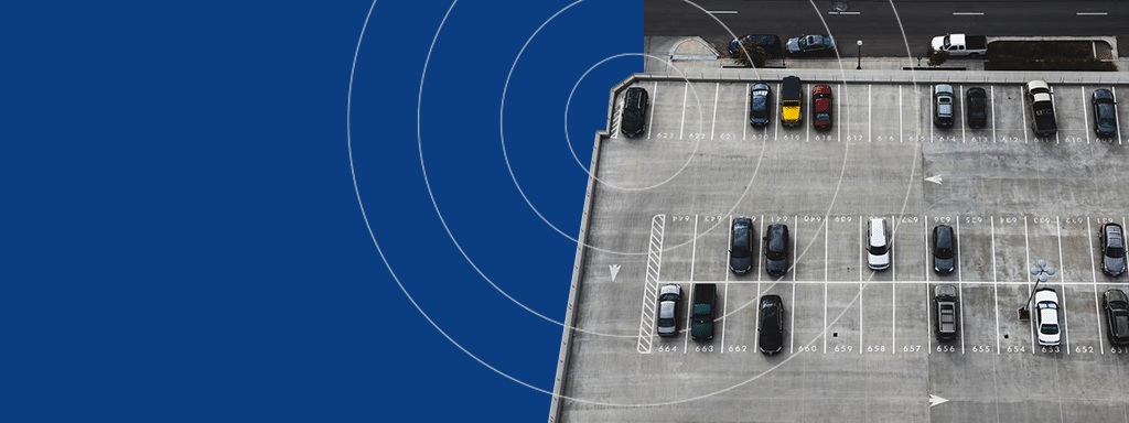 Aerial view of a parking lot with scattered vehicles, showcasing a commercial parking area. Adjacent road has more parked cars on the left. Blue overlay with concentric circles on the left side, highlighting Wayleadr's efficient parking lot management system.