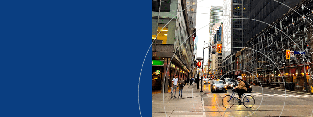 A man rides a bicycle on a city street intersection near tall buildings, navigating through pedestrians and parked cars. Construction scaffolding is visible on the right. A blue graphic related to parking management can be seen on the left side.