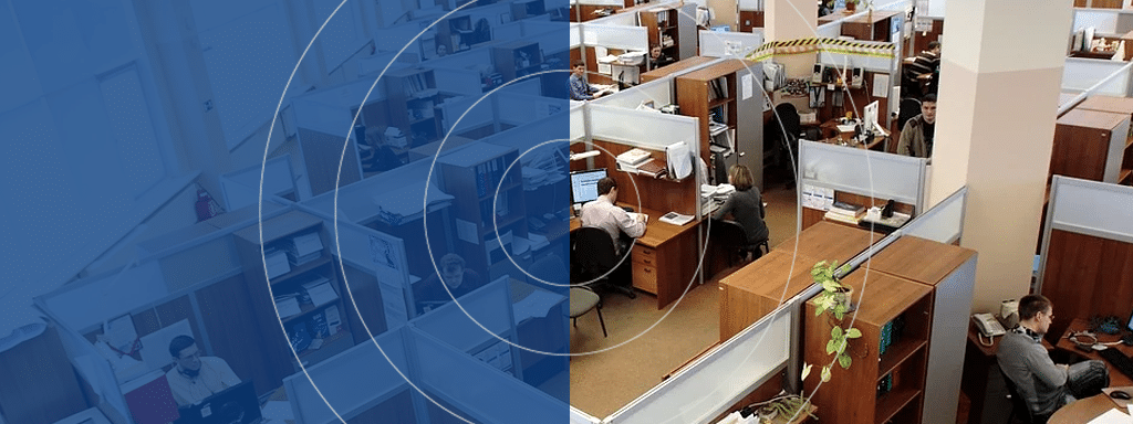 Office workspace with multiple cubicles, each occupied by employees working on computers. Some dividers have caution tape. The left part of the image features a blue overlay with concentric circles, hinting at the nearby commercial parking available for visitor parking.