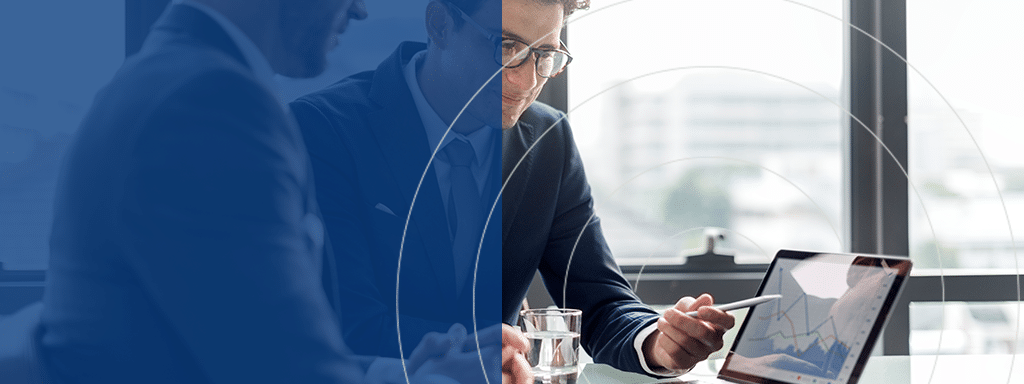 Two individuals in business attire are seated at a table, one holding a pen and looking at a tablet displaying financial charts. The background shows a blurred office environment with large windows, potentially discussing commercial parking solutions from Wayleadr.