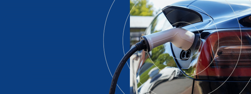 An electric car is connected to a charging station in the visitor parking area. The vehicle is plugged in, and the charging cable is visible against a blue and outdoor background.
