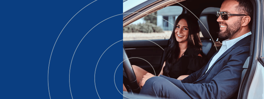 A man and woman are sitting in the front seats of a car, both smiling. The man is wearing a suit and sunglasses, and the woman is dressed in black.