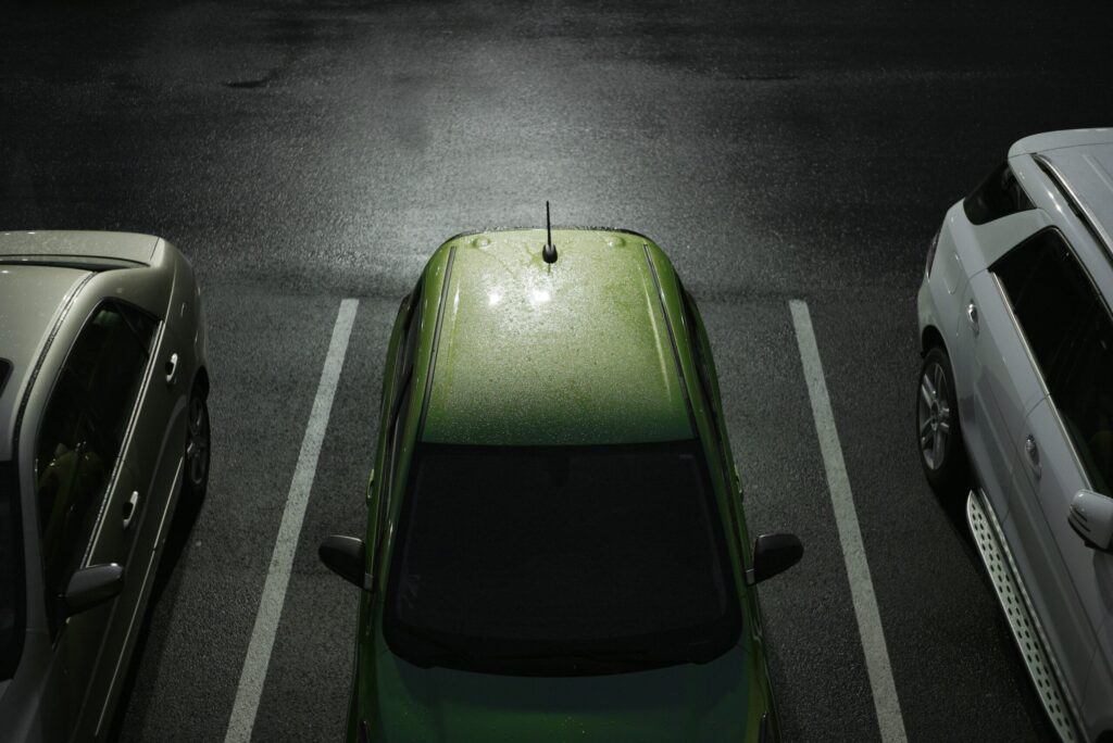A top-down view of a green car parked between a white car on the right and a silver car on the left in a dimly lit parking lot, showcasing efficient parking management.