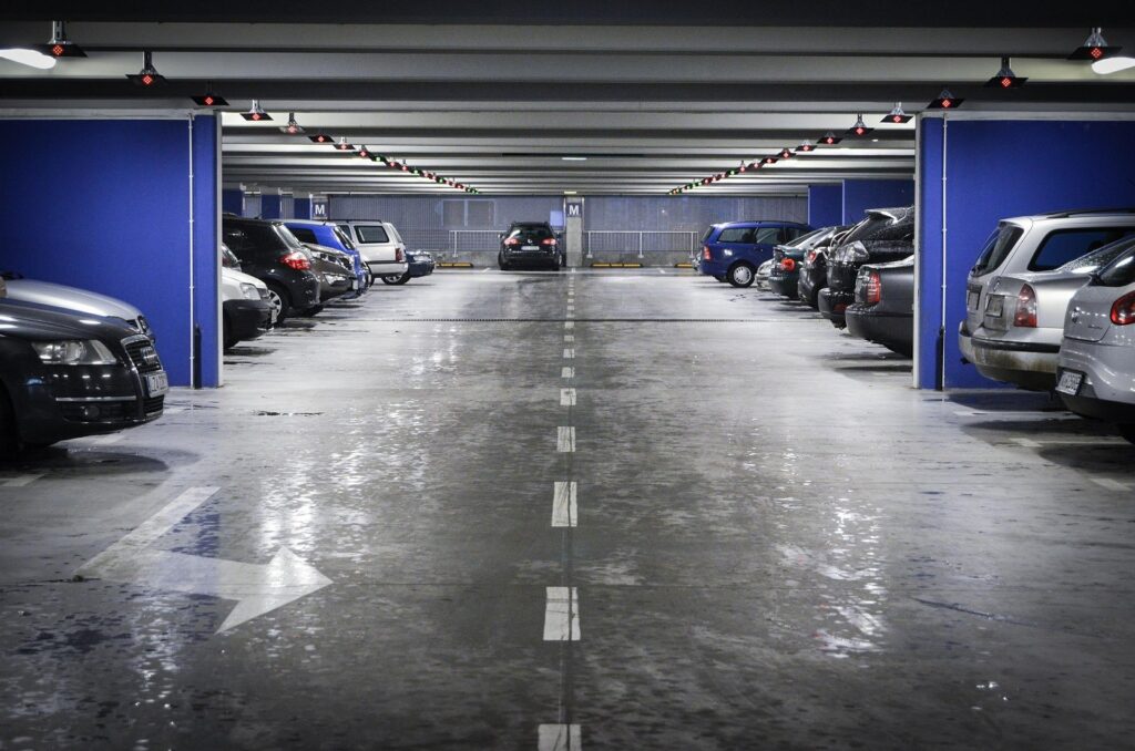 A nearly full indoor parking garage with cars parked on both sides. Blue walls and overhead lights are visible, and a single car is driving in the central lane of this commercial parking facility.