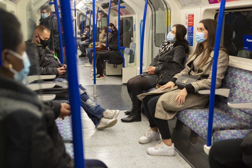 Passengers seated and standing inside a public transit train, most wearing face masks, as the train leaves the station near the commercial parking area.