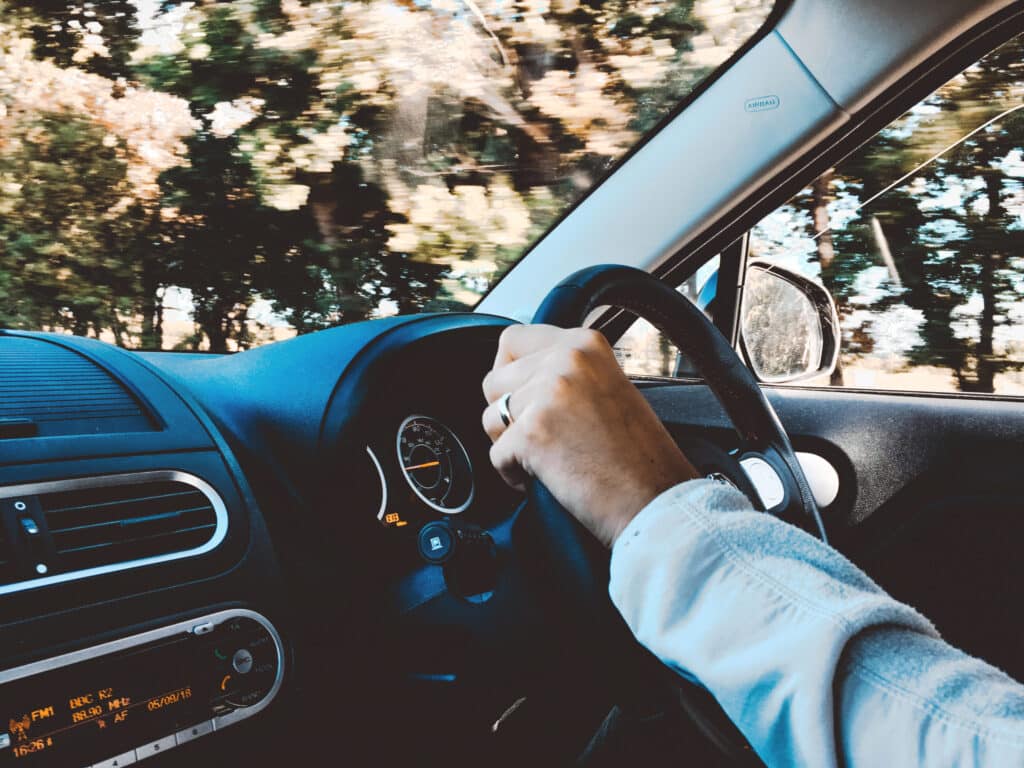 A person grips the steering wheel while driving a car with a visible dashboard and speedometer, perhaps navigating through visitor parking in a well-organized commercial parking lot. Trees are seen through the windshield.
