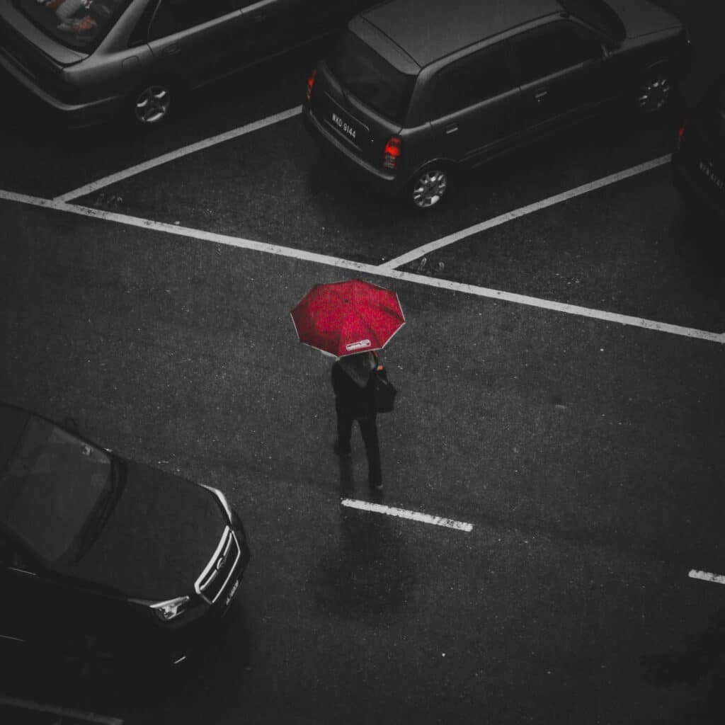 Person with a red umbrella stands on a wet road in the middle of visitor parking, surrounded by parked cars on a rainy day.