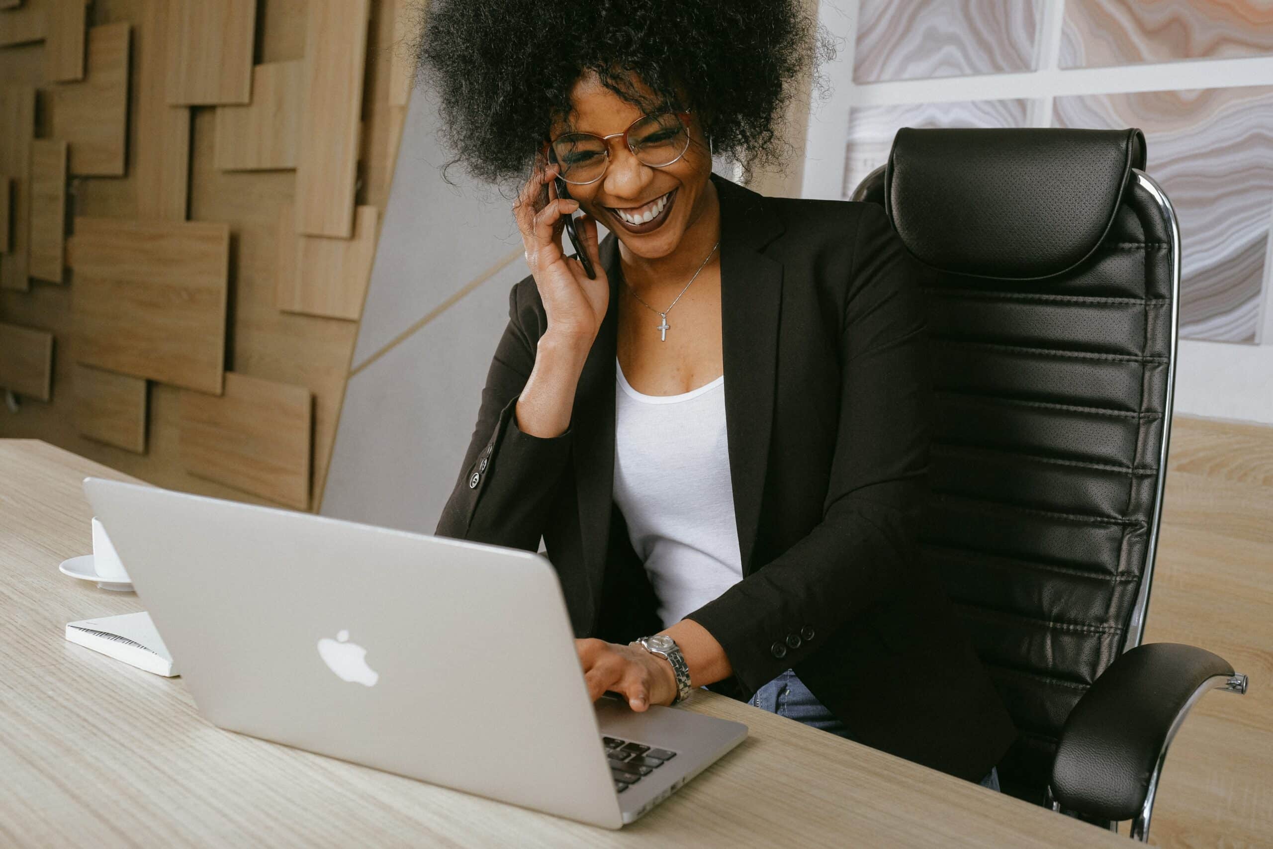 A person wearing glasses and a blazer is sitting at a desk, smiling while talking on a phone and managing visitor parking systems using a laptop.