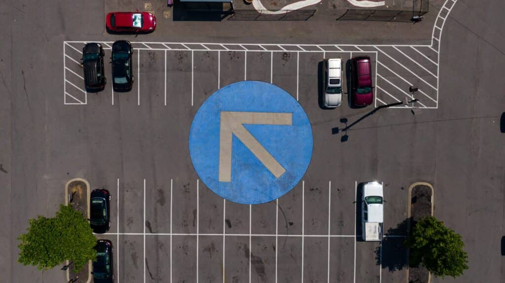 Aerial view of a parking lot with a large blue circular arrow pointing left in the center. Several cars are parked in rows on both sides of the arrow, showcasing effective parking management by Wayleadr.