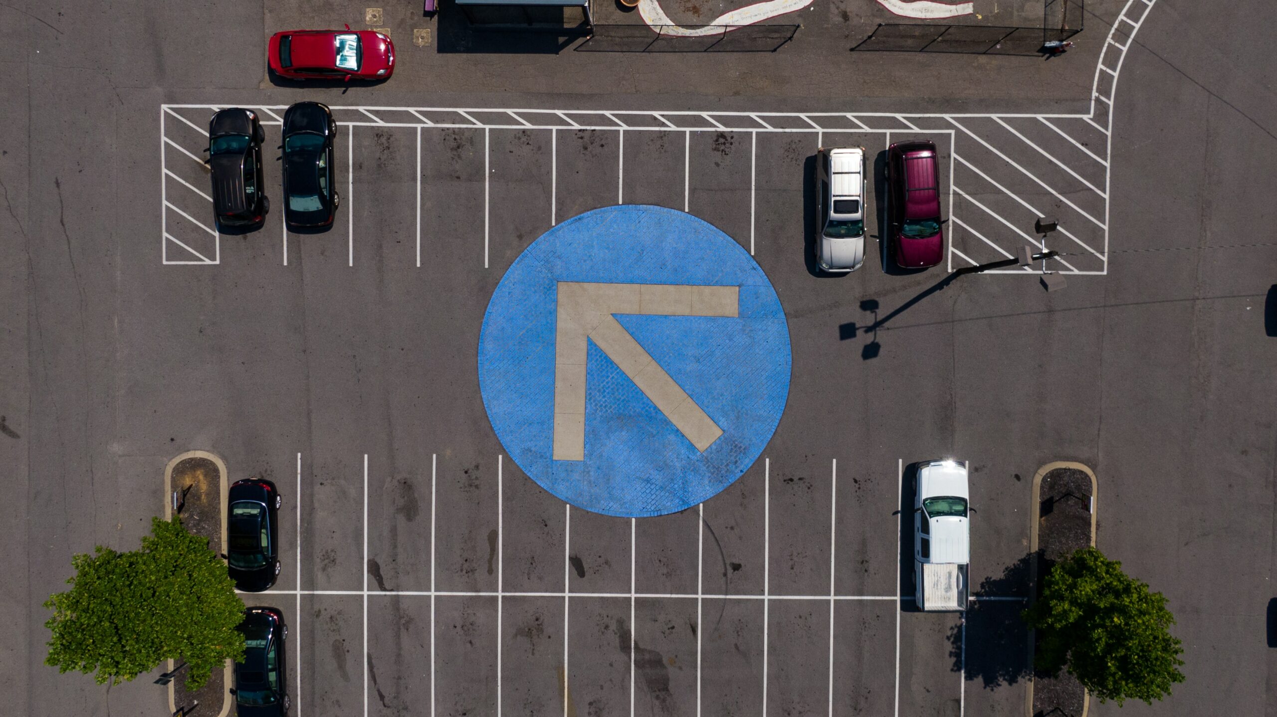 Aerial view of a parking lot with a large blue circular arrow pointing left in the center. Several cars are parked in rows on both sides of the arrow, showcasing effective parking management by Wayleadr.