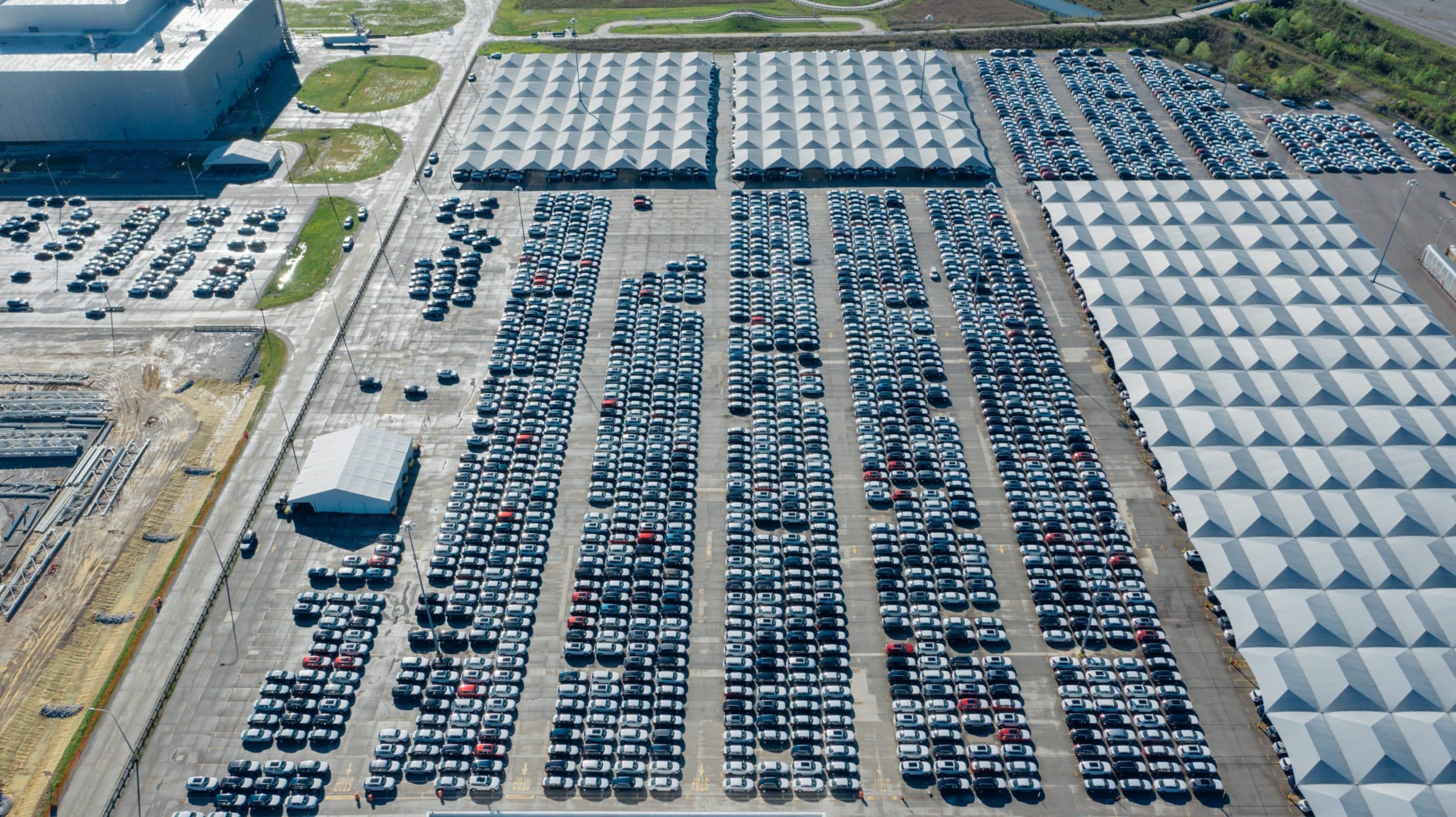 Aerial view of a large parking lot filled with rows of parked cars and several tents covering some sections, adjacent to a warehouse and surrounding grass and roads—an ideal setting for showcasing advanced parking management solutions like Wayleadr.