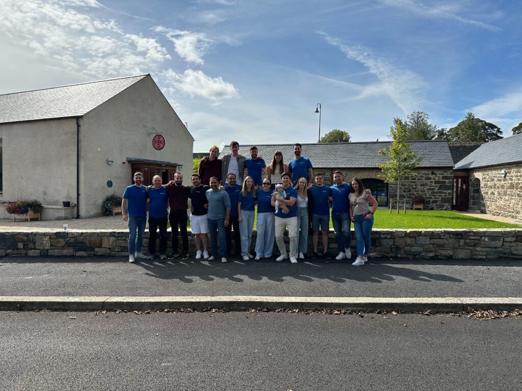 A group of 20 people, some wearing blue shirts, stand in front of a stone building with a paved road in the foreground and greenery in the background.